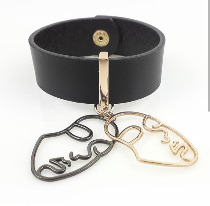 Czarny skórzany choker z metalowymi zawieszkami w kształcie konturów twarzy, jedna w kolorze grafitowym, druga złota, na białym tle.