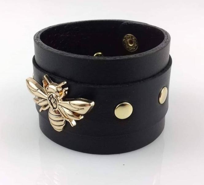 Czarna skórzana bransoletka z ozdobną złotą pszczołą i złotymi nitami, zapinana na zatrzask.