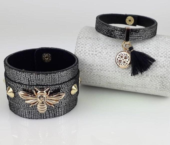 Dwa skórzane, czarno-srebrne mankiety z ozdobami: jeden z metalową pszczołą i ćwiekami, drugi z okrągłą zawieszką i czarnym chwostem, ułożone na białym tle.