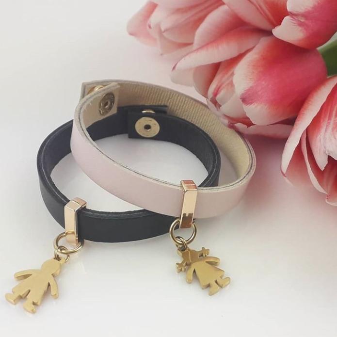 Dwa skórzane bransoletki: czarna z zawieszką chłopca i różowa z zawieszką dziewczynki, leżące na białym tle z rozmytymi różowymi tulipanami w tle.