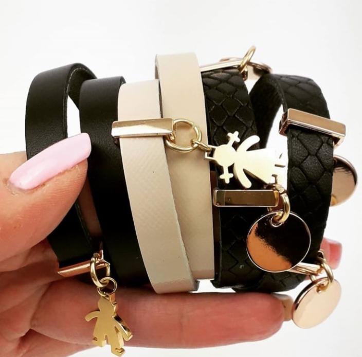 Ręka trzymająca zestaw kilku bransoletek: czarna skórzana, beżowa i czarna pleciona, z zawieszkami w kształcie ludzików i kółek w kolorze złota.
