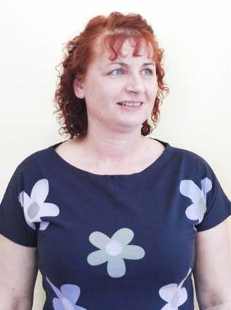 Portret uśmiechniętej kobiety z rudymi kręconymi włosami, ubrana w granatową koszulkę w kwiaty na jasnym tle.