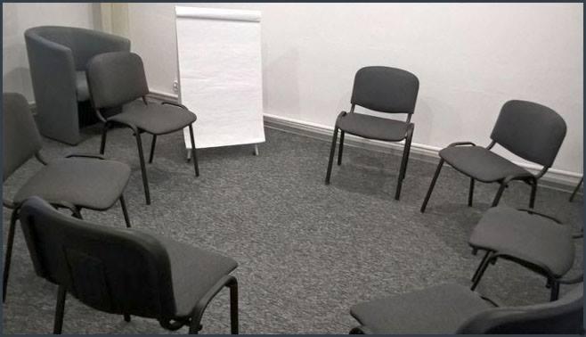 Krzesła ustawione w okręgu w pokoju z szarym dywanem i tablicą flipchart, przygotowane do sesji grupowej.