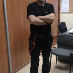 Mężczyzna w czarnym ubraniu roboczym z logo firmy stojący na tle szafy przesuwnej i biurka z krzesłem.