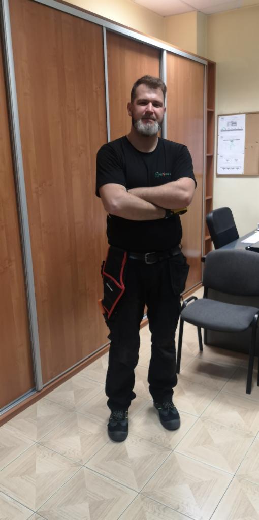 Mężczyzna w czarnym ubraniu roboczym z logo firmy stojący na tle szafy przesuwnej i biurka z krzesłem.