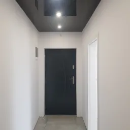 Korytarz po remoncie: czarne, błyszczące sufity napinane z trzema punktami oświetleniowymi, białe ściany i szara podłoga z płytek, widoczne drzwi wejściowe i ościeżnica drzwi wewnętrznych.