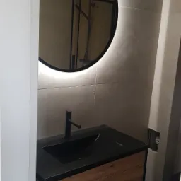 Nowoczesna łazienka z okrągłym lustrem z podświetleniem LED, czarną umywalką nablatową na szafce z drewnianym frontem i czarnym wykończeniem oraz widoczną kabiną prysznicową w odbiciu lustra.