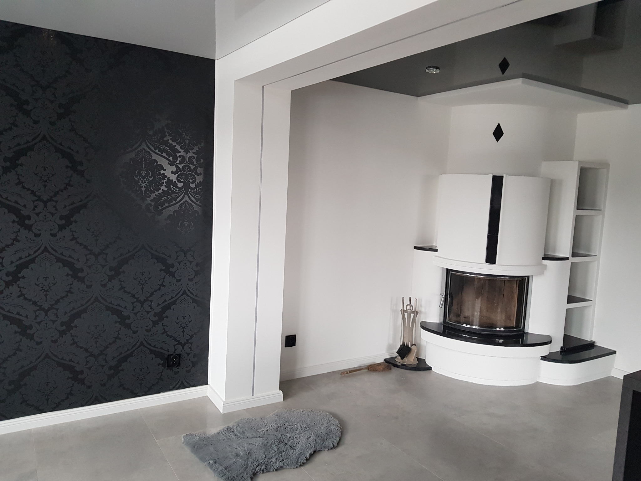 Eleganckie wnętrze z kominkiem i czarną tapetą w ornamenty, szary dywanik na podłodze, białe ściany i sufit z czarnymi elementami dekoracyjnymi.