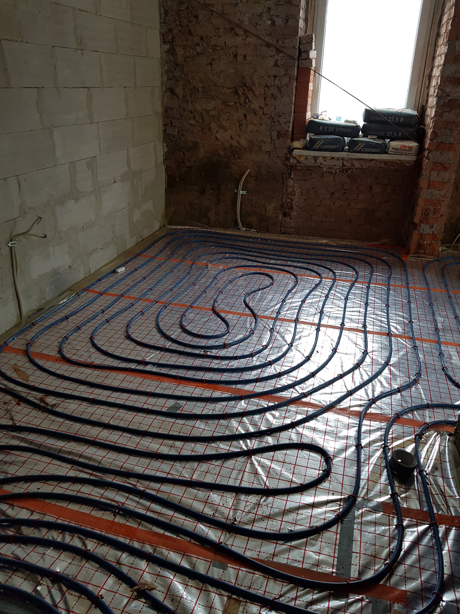 Instalacja ogrzewania podłogowego z rur w surowym pomieszczeniu, widoczne worki z cementem przy oknie.