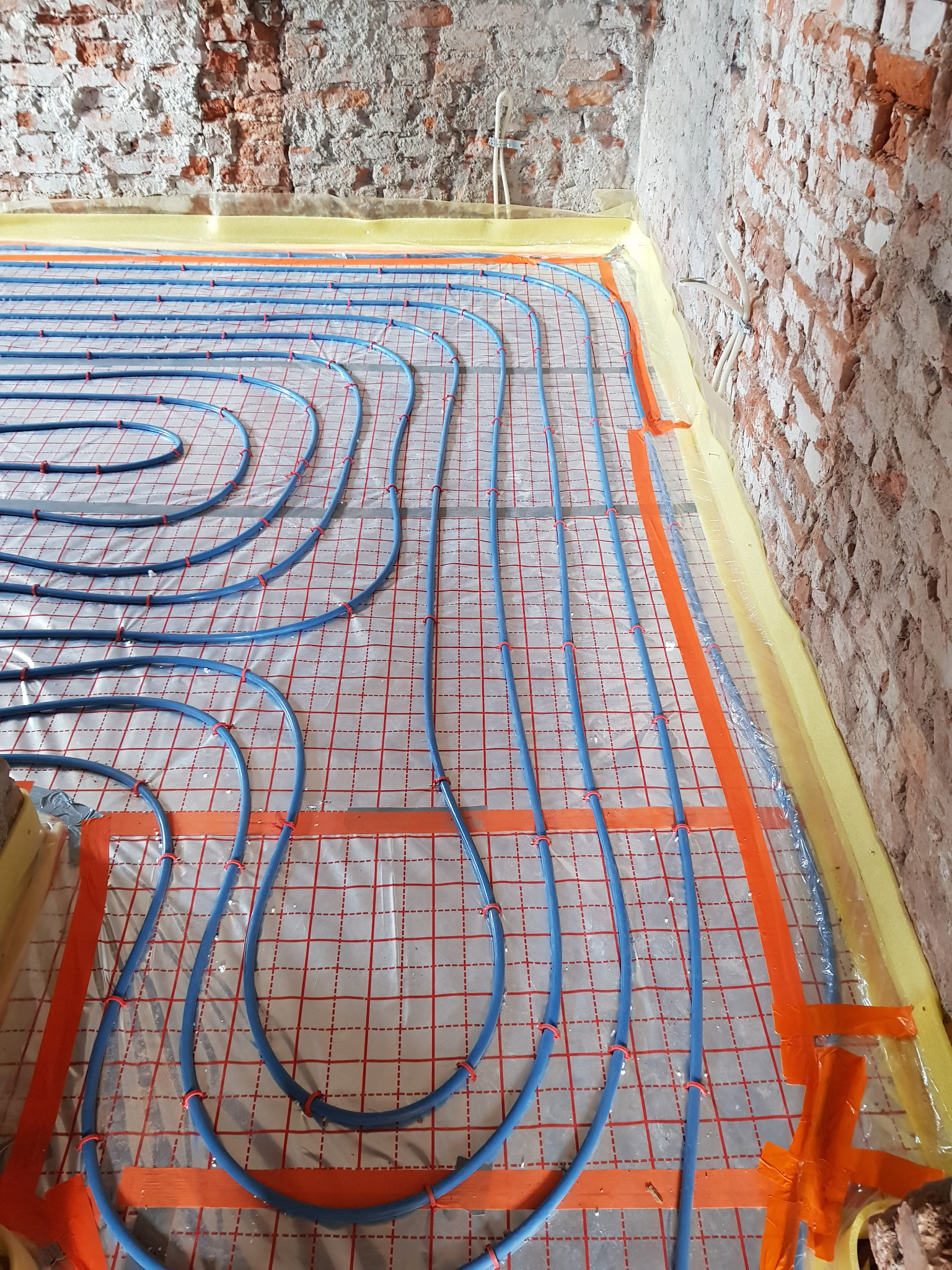 Montaż ogrzewania podłogowego w Kamiennej Górze: niebieskie rury ułożone na folii z czerwoną siatką, widoczne surowe, ceglane ściany w tle.