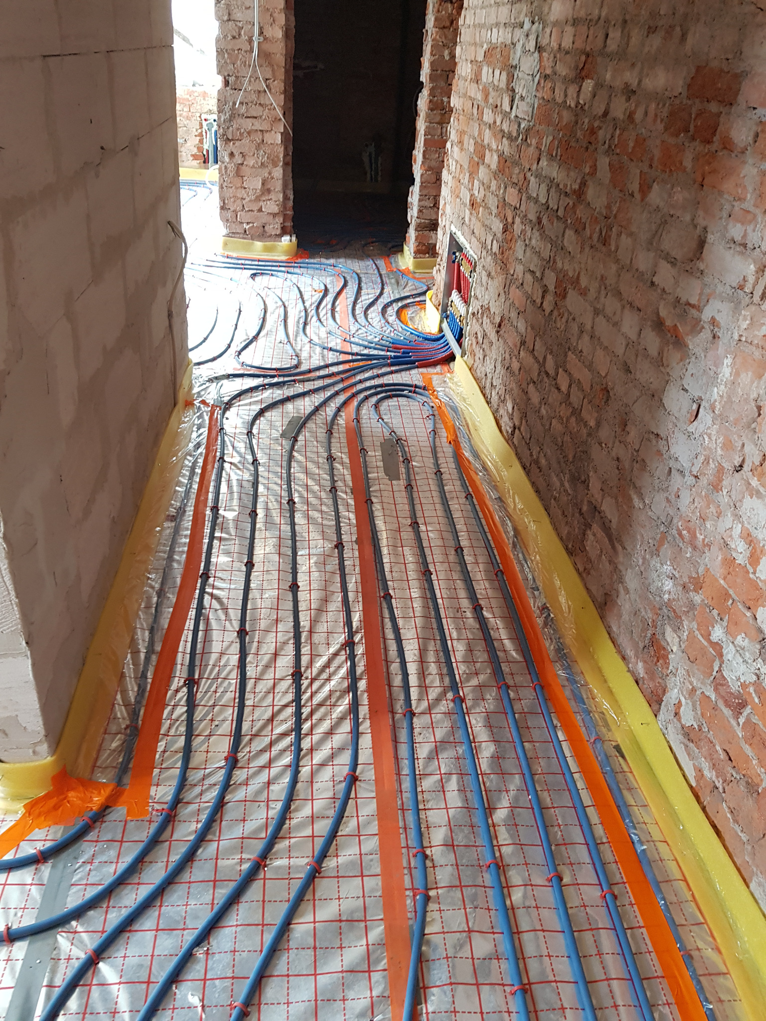 Instalacja ogrzewania podłogowego w pomieszczeniu z surowymi ścianami z cegły i tynkowanymi. Niebieskie rury układane na folii izolacyjnej z czerwoną siatką, mocowane pomarańczowymi taśmami. W tle...