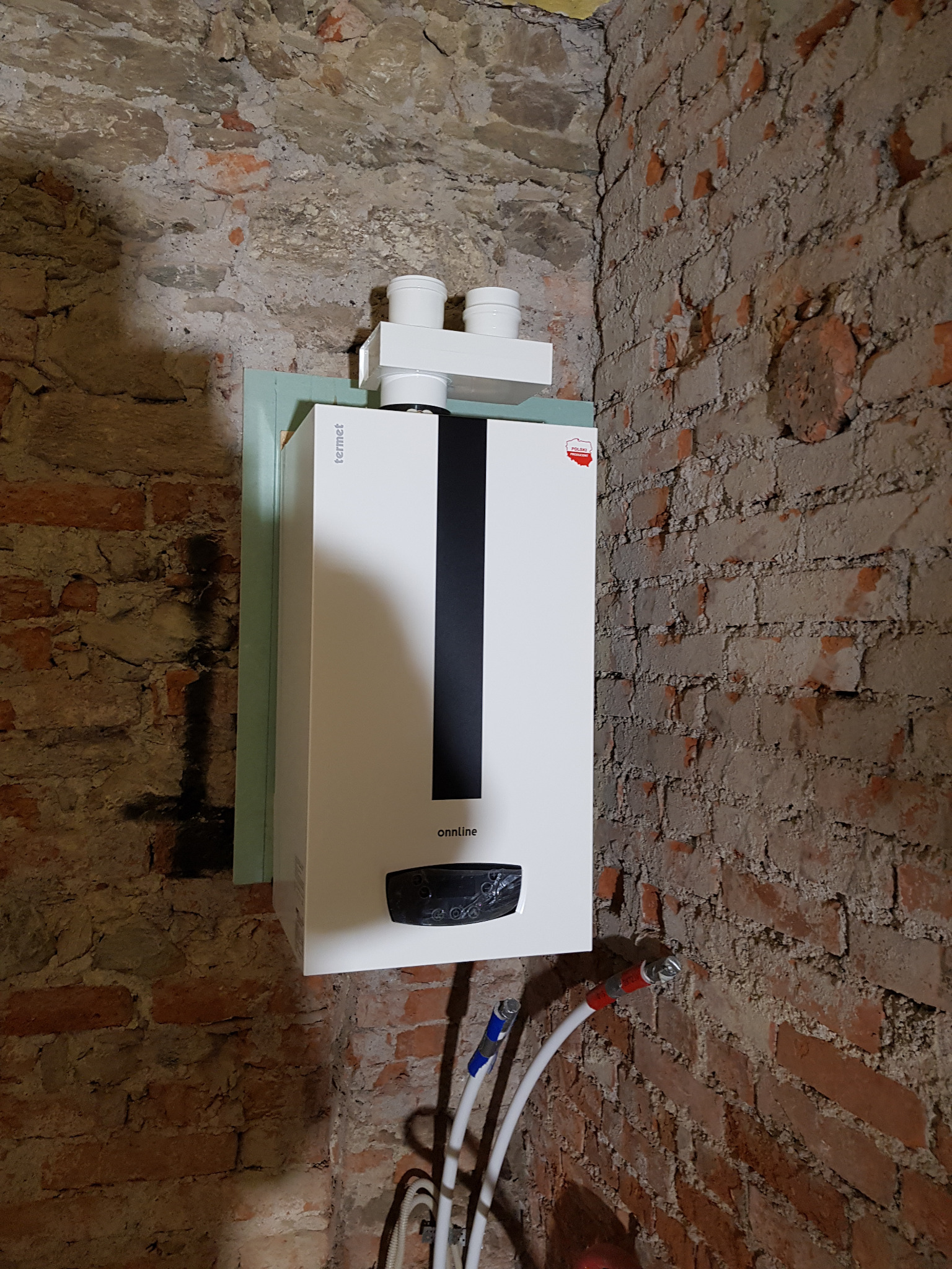 Nowoczesny, biały piec gazowy marki Termet, model Onnline, zamontowany na ścianie z surowej, ceglanej faktury, z widocznymi rurami przyłączeniowymi.