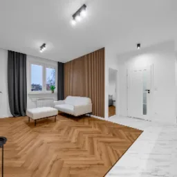 Jasne, nowocześnie urządzone wnętrze mieszkania z parkietem w jodełkę, ścianą z drewnianych lameli, białym narożnikiem i marmurową podłogą przechodzącą w parkiet.