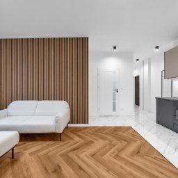 Agencja Remontowa - Nowoczesne wnętrze: parkiet w jodełkę, ściana z drewnianych lameli, biała sofa i marmurowe płytki. Minimalistyczna kuchnia w tle.