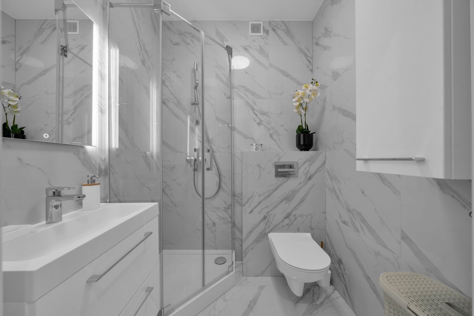 Elegancka łazienka z marmurowymi płytkami, kabiną prysznicową ze szklanymi drzwiami, wiszącą toaletą, białą umywalką z szafką i dekoracyjną orchideą w czarnym wazonie.