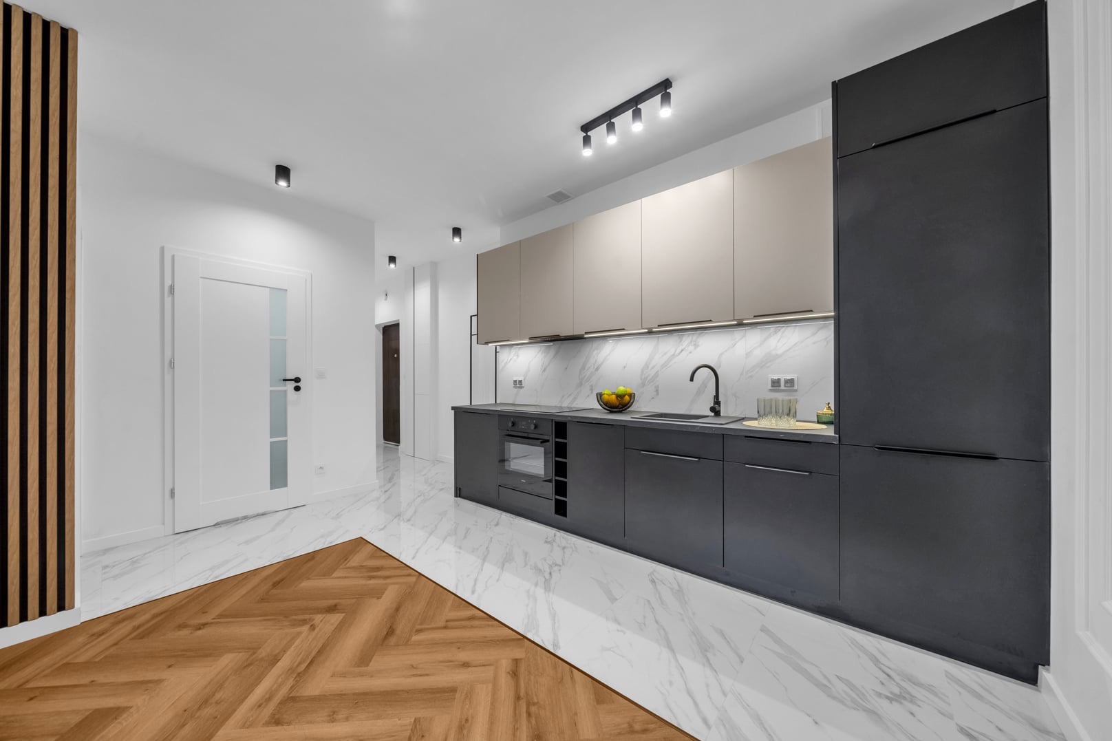 Nowoczesna kuchnia z szaro-beżowymi szafkami, marmurowym blatem i podświetleniem LED, drewniana podłoga w jodełkę przechodzi w marmurowe płytki, widoczny fragment drewnianej ścianki działowej.