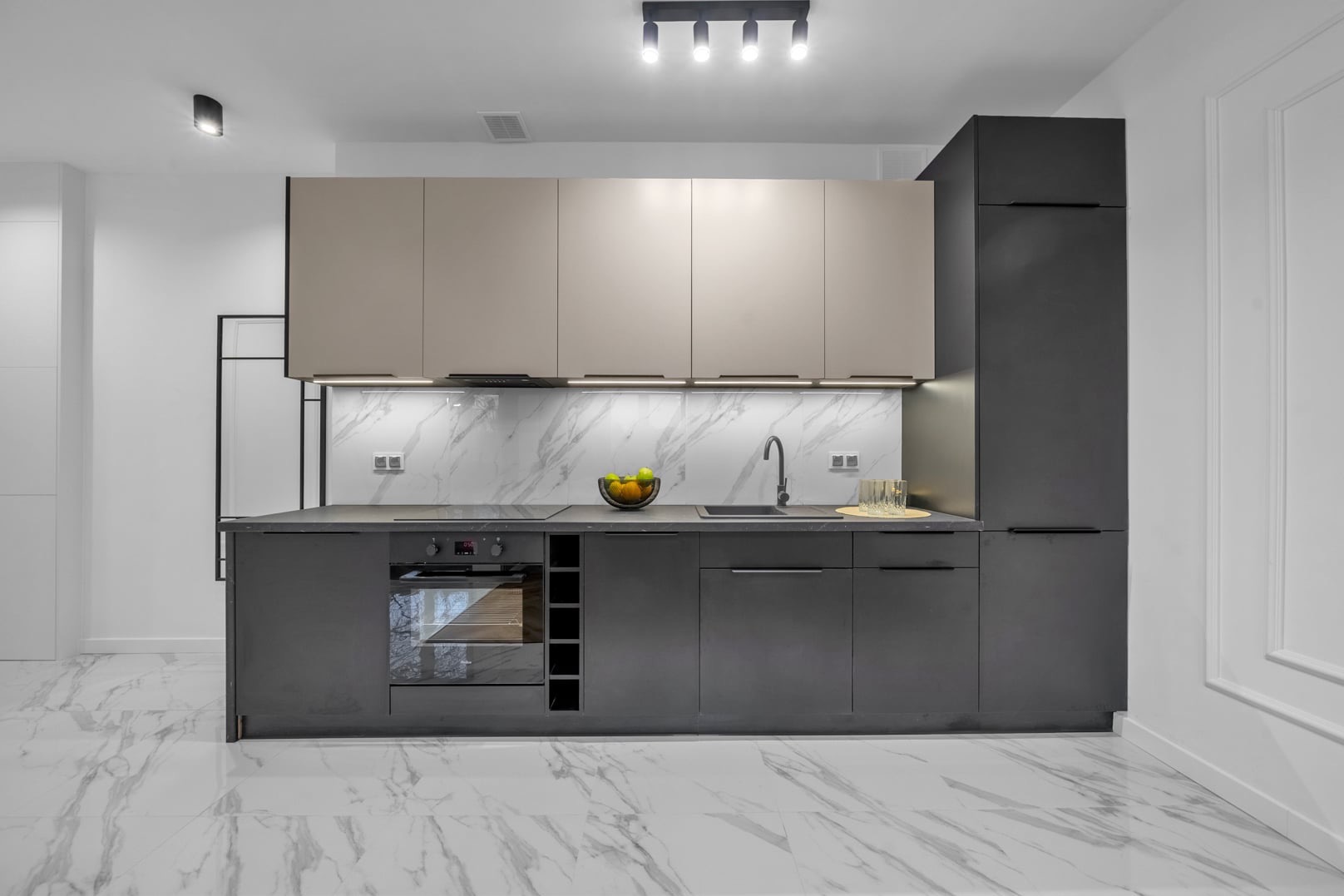 Nowoczesna kuchnia z szarymi szafkami dolnymi i beżowymi górnymi, marmurowym backsplashiem podświetlonym LED, armaturą w kolorze grafitu oraz podłogą z białego marmuru z szarymi żyłami.