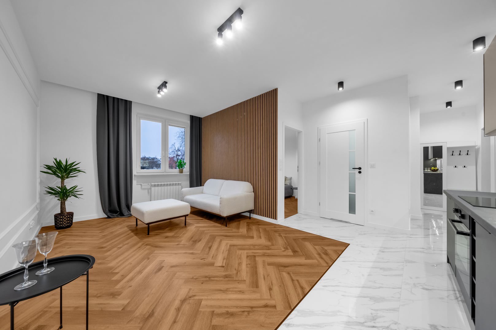 Jasne, nowocześnie urządzone wnętrze mieszkania z parkietem w jodełkę, ścianą z drewnianych lameli, białym narożnikiem i marmurową podłogą przechodzącą w parkiet.