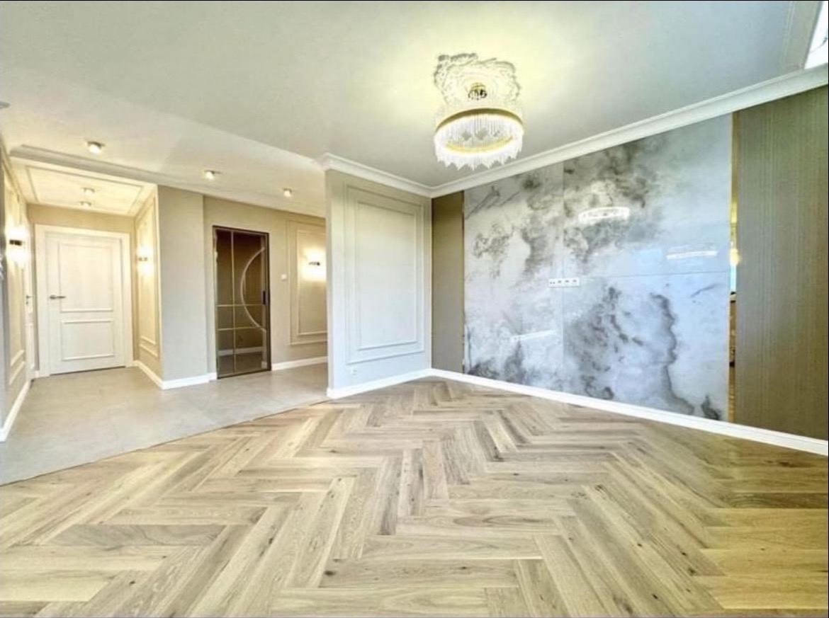 Eleganckie wykończenie wnętrza: parkiet w jodełkę, marmurowa ściana, sztukateria i designerski żyrandol. Widok na korytarz z drzwiami.