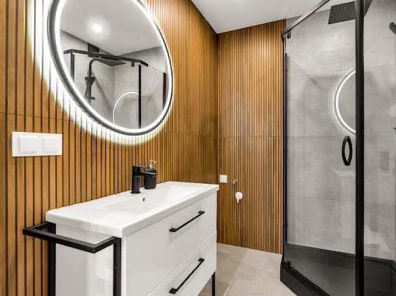 Nowoczesna łazienka z drewnianymi panelami ściennymi, białą szafką z umywalką i podświetlanym lustrem. Kabina prysznicowa z czarnymi ramami.