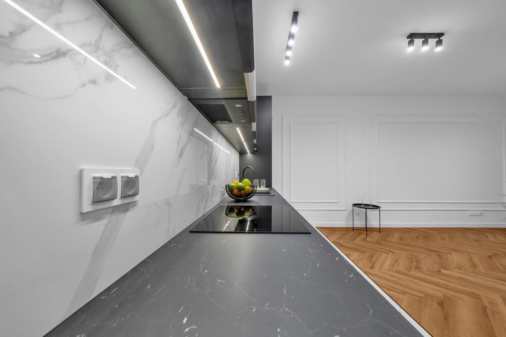 Nowoczesna kuchnia z marmurową ścianą, czarnym blatem i oświetleniem LED. Na blacie misa z owocami. Parkiet w jodełkę. Minimalistyczny design.