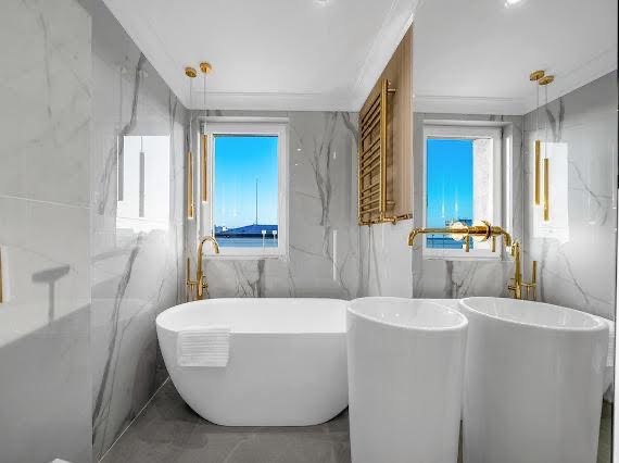 Elegancka łazienka z marmurowymi ścianami, wolnostojącą wanną z ręcznikiem, złotą armaturą i dwoma umywalkami. Widok na zewnątrz przez okno.