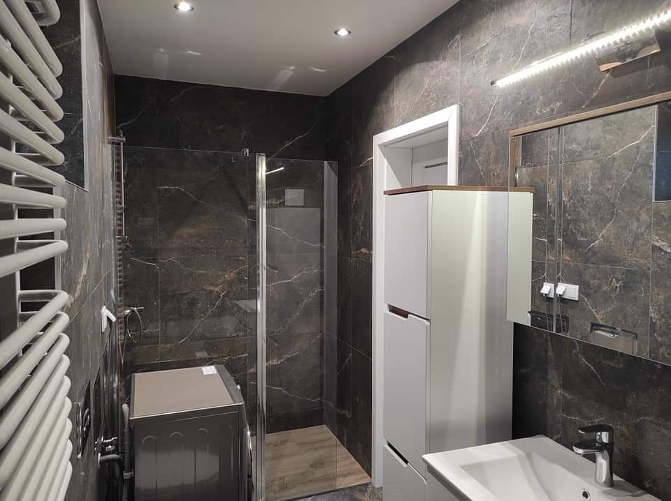 Nowoczesna łazienka z ciemnymi płytkami imitującymi marmur, kabiną prysznicową typu walk-in, białą szafką łazienkową i pralką w rogu.