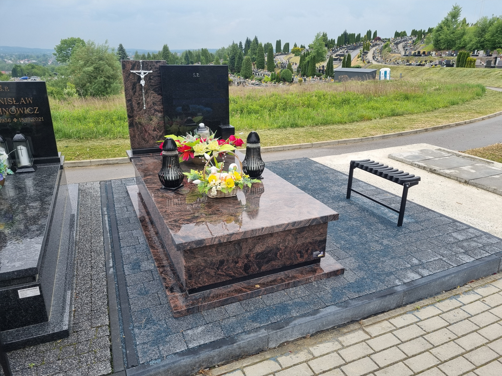 Granitowy pomnik z ciemnobrązowego kamienia Aurora, zdobiony kwiatami i lampionami, na tle cmentarza z rozległym widokiem na okolicę.