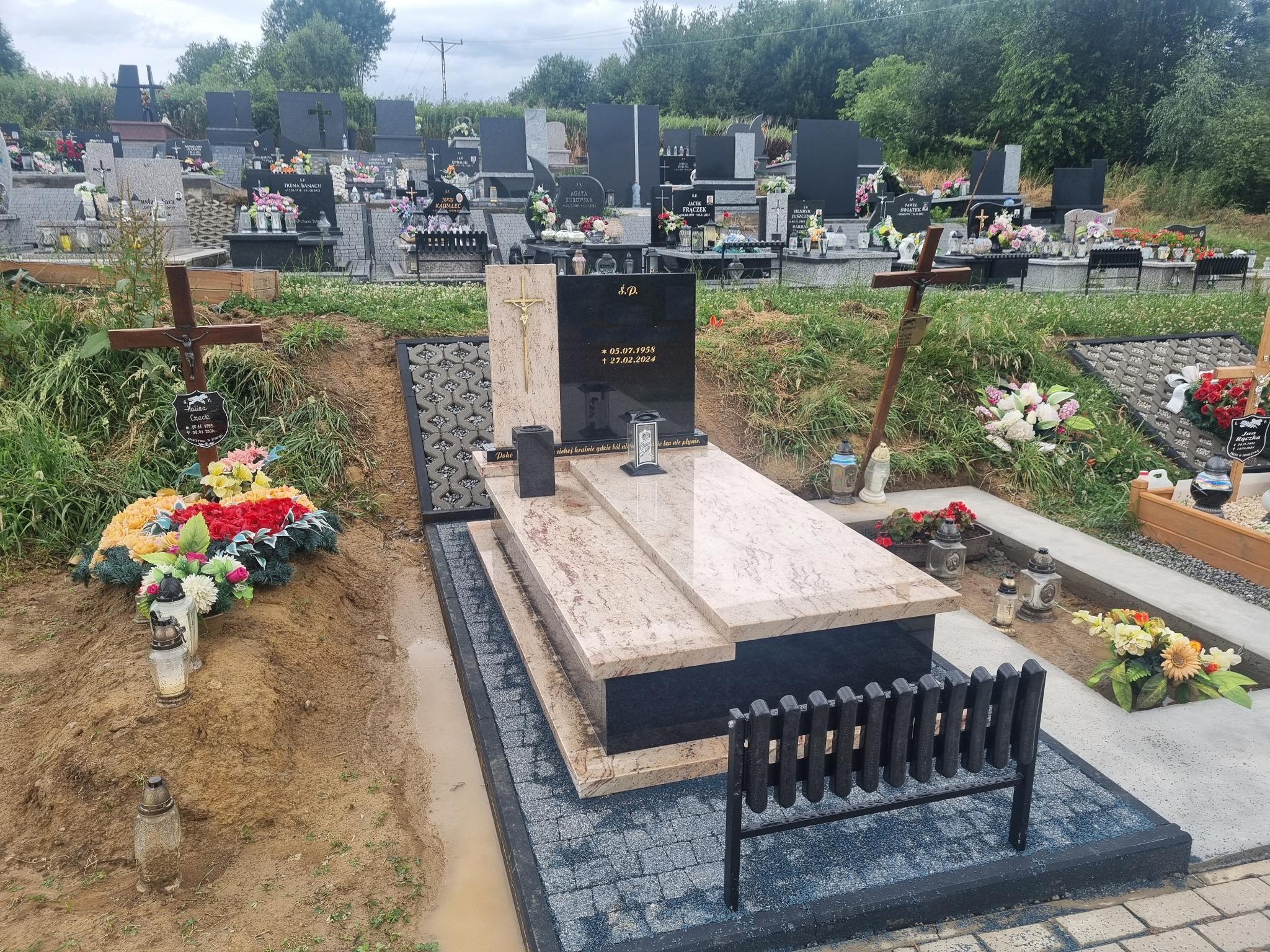 Granitowy nagrobek ziemny Shivakashi i Bengal Black na cmentarzu, otoczony kwiatami i zniczami, z widocznym fragmentem kostki brukowej i metalowej ławki.