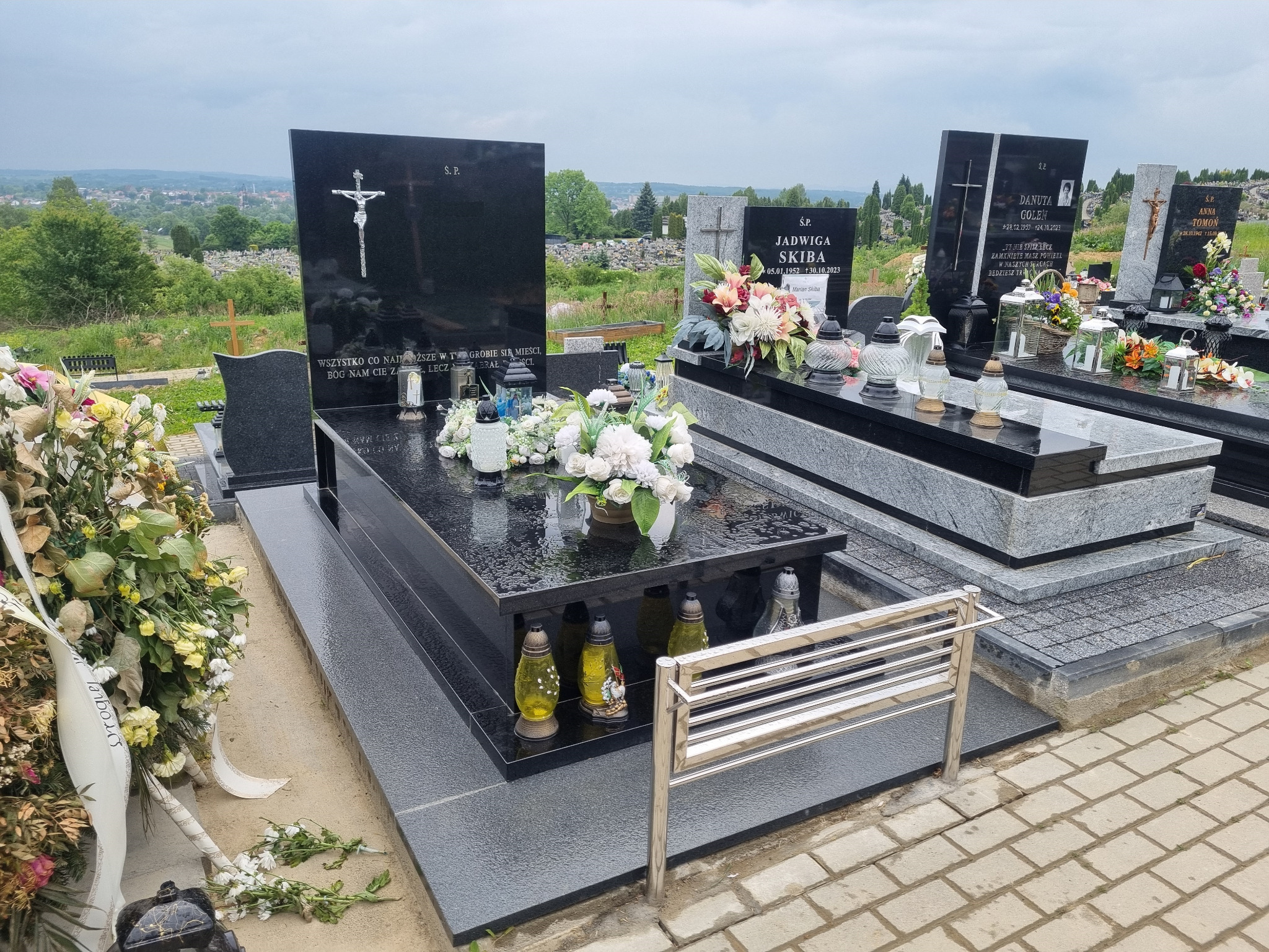 Czarny granitowy pomnik z krzyżem i srebrną ławeczką na cmentarzu, w tle widok na miasto.