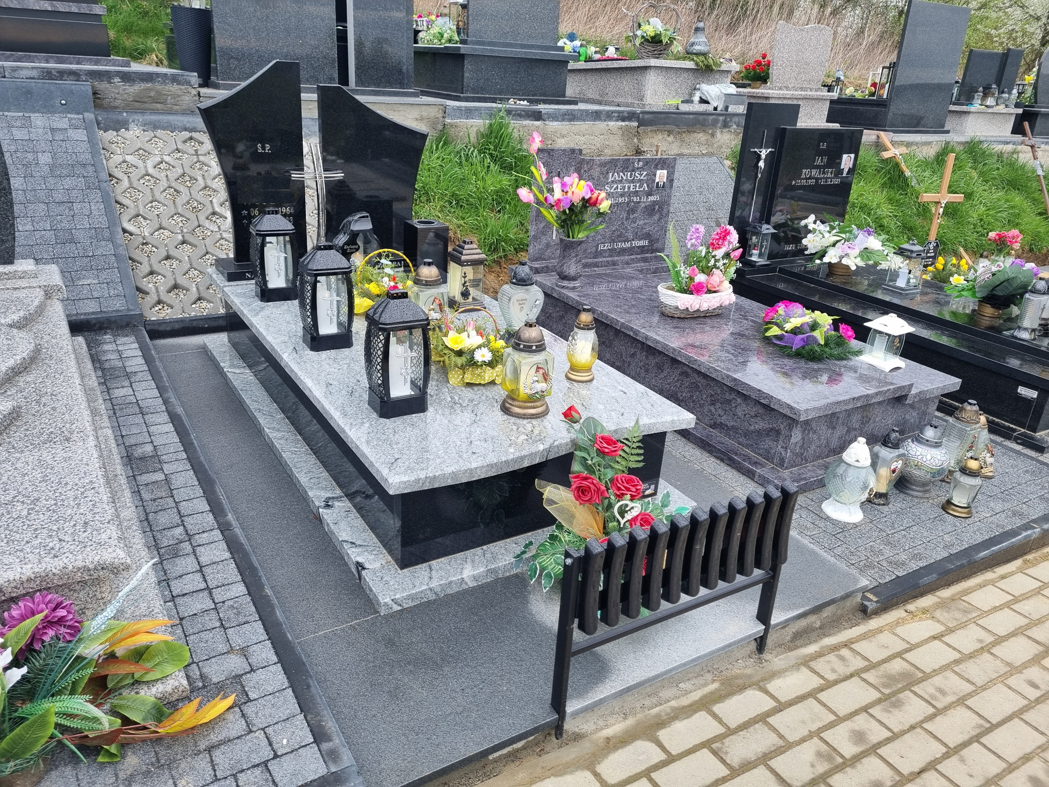 Wielopoziomowy pomnik z jasnego i ciemnego granitu, ozdobiony stalowym krzyżem, kwiatami w wazonach i lampionami, na tle innych grobów.