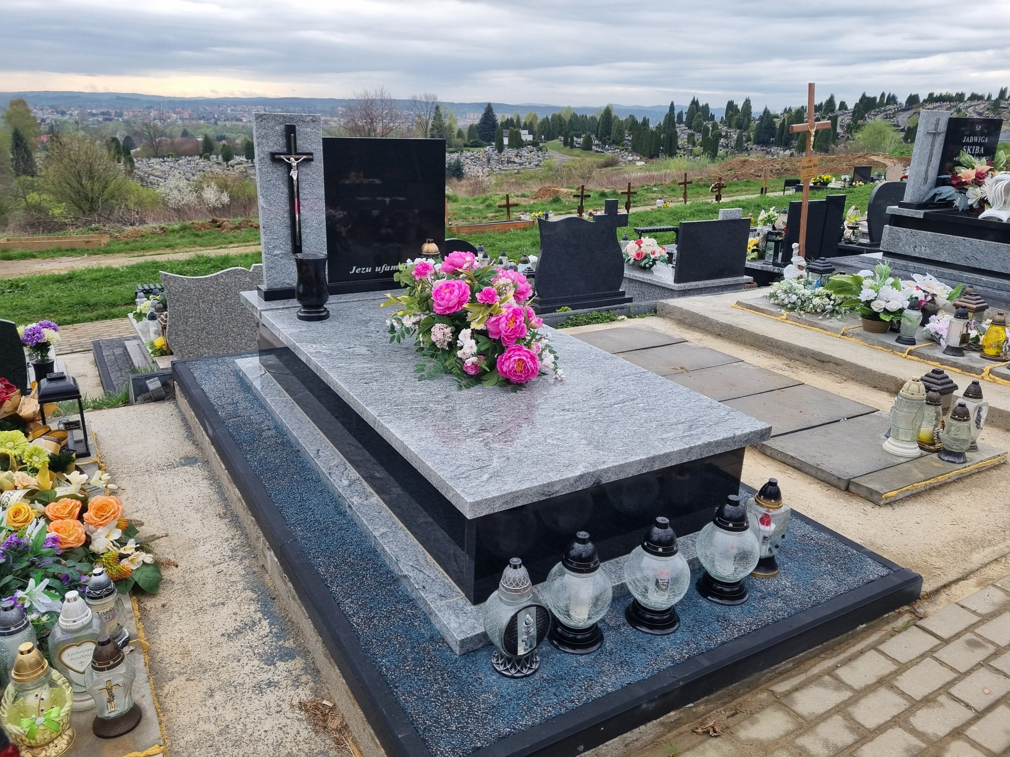 Dwukolorowy, granitowy pomnik z czarną płytą nagrobkową i bukietem różowych kwiatów, otoczony lampionami na tle cmentarza z widokiem na miasto.