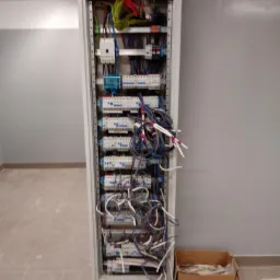 Otwarta szafa rozdzielcza z licznymi zabezpieczeniami nadprądowymi i przewodami, częściowo uporządkowanymi, z kartonowym pudłem z odpadami po instalacji obok, na tle szarej ściany.