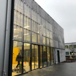 Budynek z nowoczesną fasadą, widoczne duże przeszklenia i metalowa perforowana osłona. Wewnątrz budynku osoba w kurtce, prawdopodobnie podczas prac montażowych.