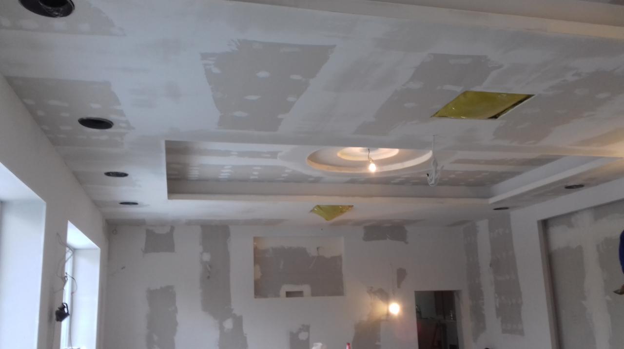 Wnętrze w trakcie prac wykończeniowych: sufit z dekoracyjnymi elementami i otworami na oświetlenie, ściany częściowo pokryte gładzią, przygotowane do malowania.