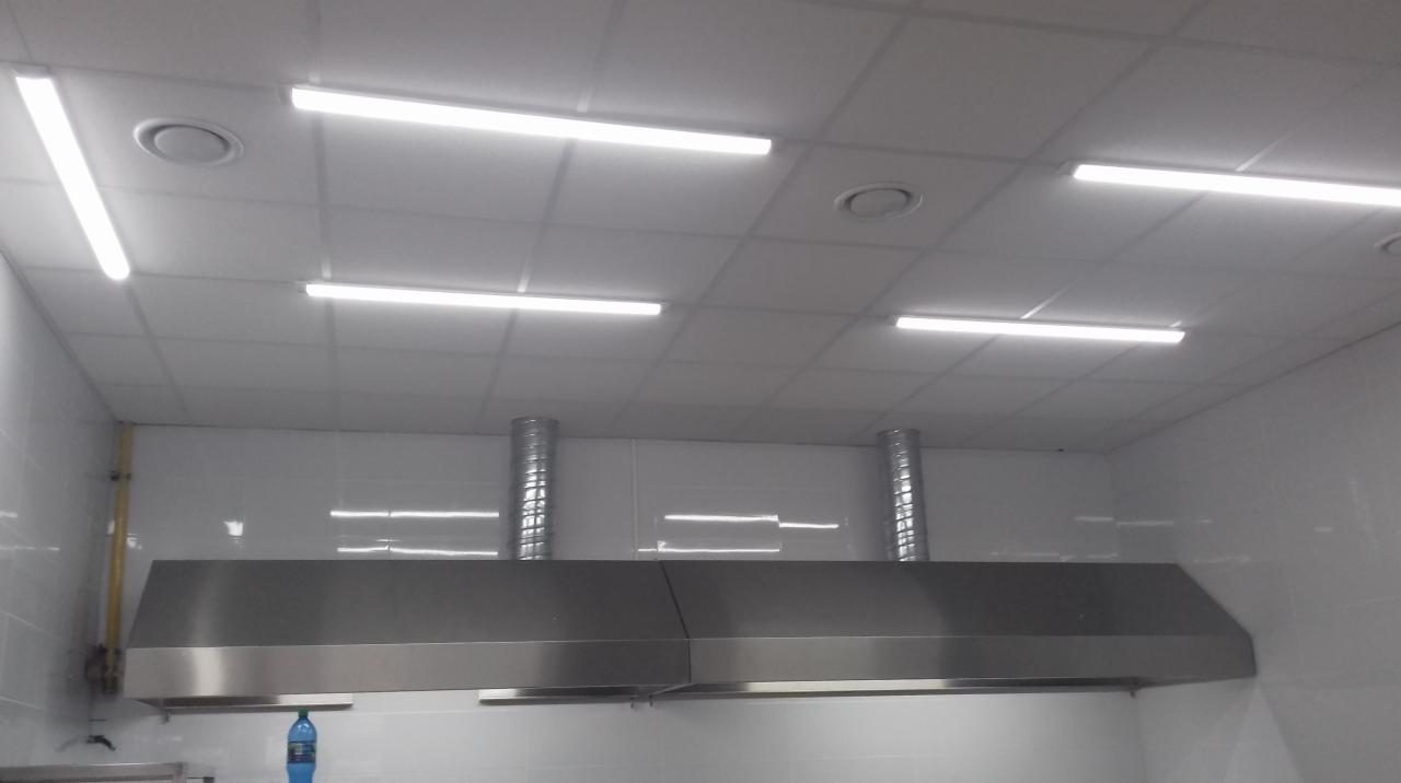 Wnętrze kuchni przemysłowej z zamontowanym okapem ze stali nierdzewnej i białymi kafelkami na ścianach oraz oświetleniem jarzeniowym na suficie kasetonowym.