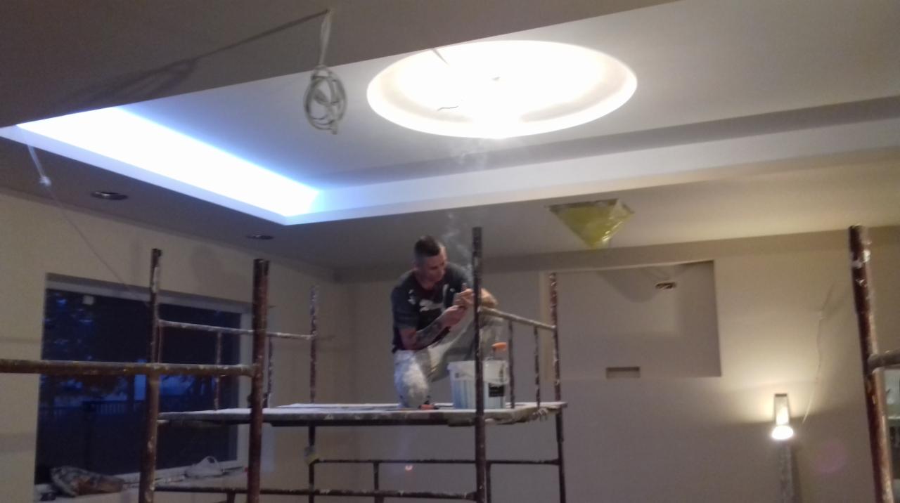 Mężczyzna na rusztowaniu szpachluje sufit z oświetleniem LED w pomieszczeniu podczas remontu.