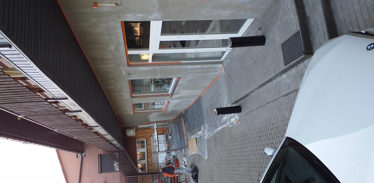 Elewacja budynku w trakcie prac malarskich, zabezpieczone okna taśmą malarską, widoczne narzędzia i materiały budowlane, obok zaparkowany biały samochód.