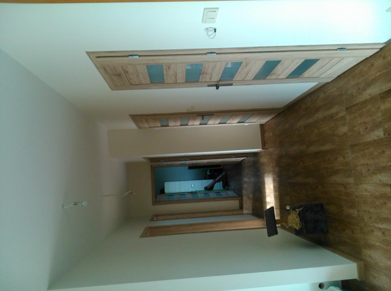 Wnętrze korytarza z drewnianymi drzwiami z przeszkleniami, świeżo pomalowane ściany w odcieniach bieli i brązowa podłoga imitująca drewno, widok w głąb mieszkania.