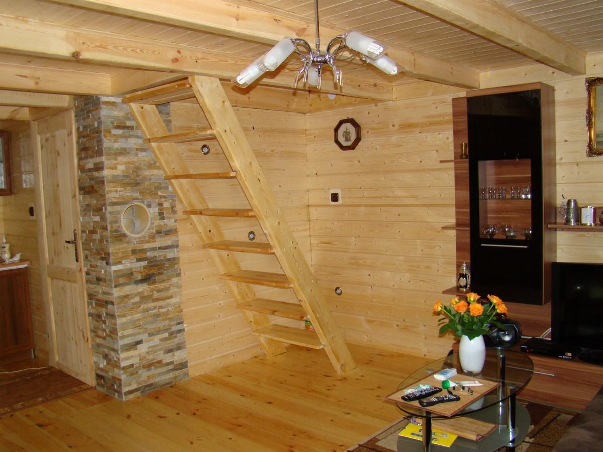 Wnętrze domku drewnianego 50m2 z drewnianymi schodami bez poręczy prowadzącymi na antresolę, ściany wyłożone boazerią, obok schodów dekoracyjny filar z kamienia, na podłodze parkiet, w tle meble...