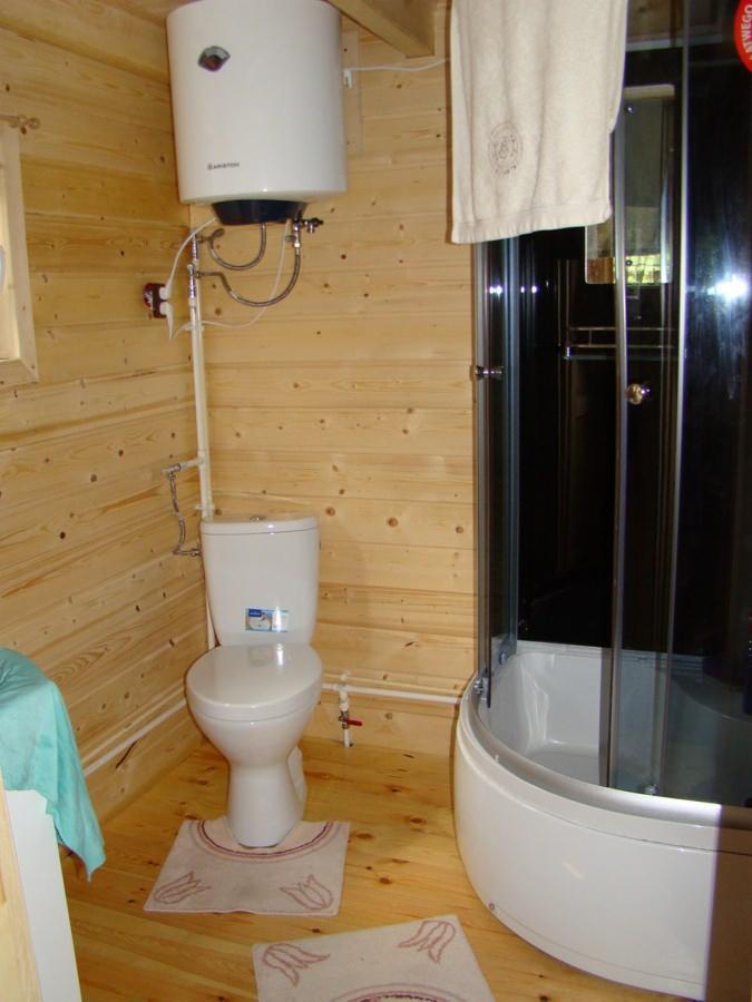 Wnętrze łazienki w domku drewnianym 50m2: kabina prysznicowa, toaleta i bojler na tle drewnianych ścian i podłogi, z dywanikami w tulipany.