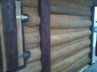 Ściana zewnętrzna drewnianego domku o powierzchni 50m2, obita imitacją bala w kolorze jasnego drewna, z widoczną brązową ramą okienną i metalowymi zawiasami w kolorze zielonym.