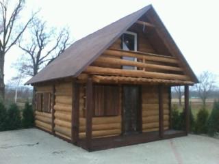 Mały drewniany domek letniskowy obity imitacją bala, z brązowym dachem dwuspadowym i niewielkim balkonem na piętrze. Widoczne drewniane okiennice i zadaszony taras.