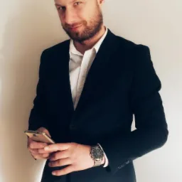 Mężczyzna w garniturze, z brodą, trzymający złoty smartfon w dłoniach, na nadgarstku widoczny zegarek z metalową bransoletą.