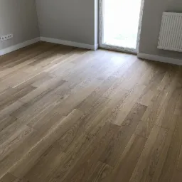 Jasne, lakierowane deski parkietowe ułożone w nowym pokoju z grzejnikiem i listwami przypodłogowymi, widoczne gniazdka elektryczne po lewej stronie.