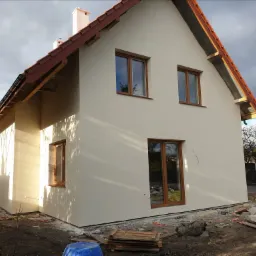 Nowo otynkowana elewacja domu jednorodzinnego z brązowymi oknami i dachem z czerwonej dachówki, widoczne materiały budowlane i niezagospodarowany teren wokół.