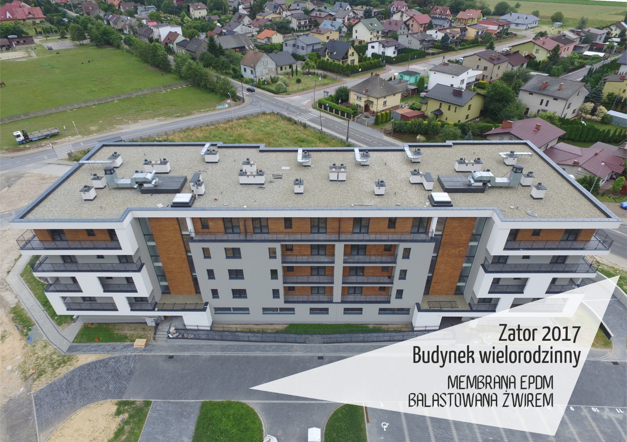 Widok z góry na dach wielorodzinnego budynku z membraną EPDM balastowaną żwirem, liczne kominy wentylacyjne, w tle zabudowa jednorodzinna.