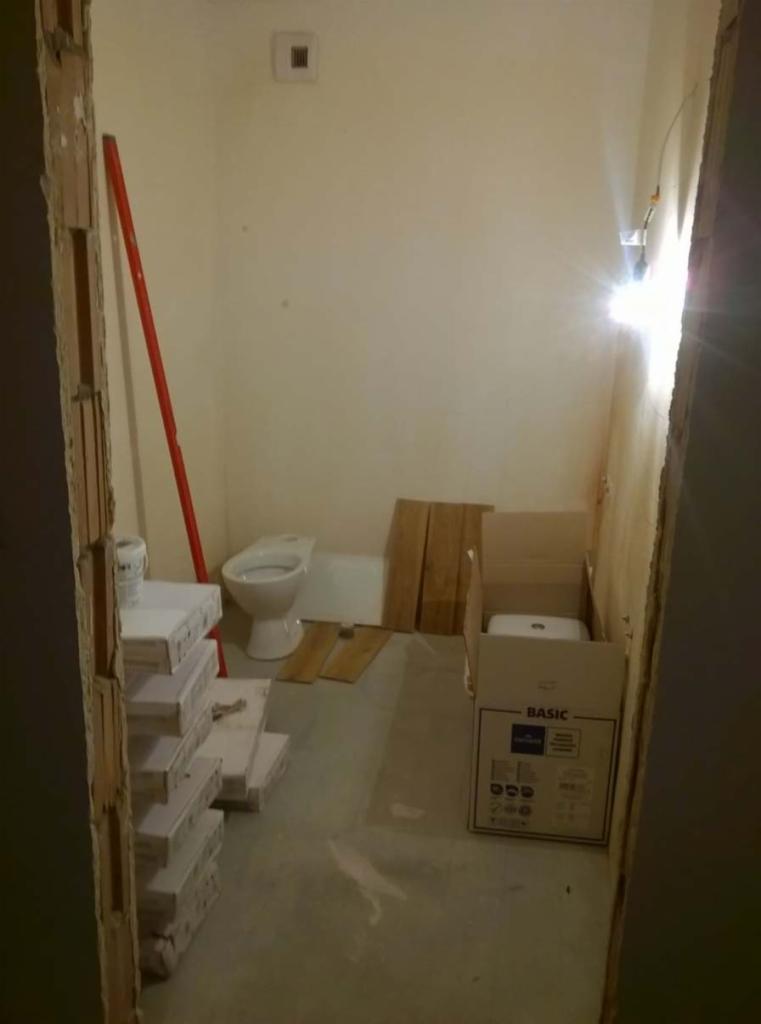 Łazienka w trakcie remontu: widoczny sedes, płytki w opakowaniach, deski podłogowe i pudełko z umywalką, czerwona poziomica oparta o ścianę.