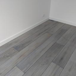 Usługi Parkieciarskie - Szare panele podłogowe ułożone w rogu pokoju z białymi ścianami i listwami przypodłogowymi.