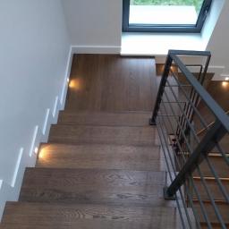 Usługi Parkieciarskie - Nowoczesne schody drewniane z metalową balustradą, oświetleniem punktowym w ścianie i pod stopniami, widok z góry na pierwsze stopnie.